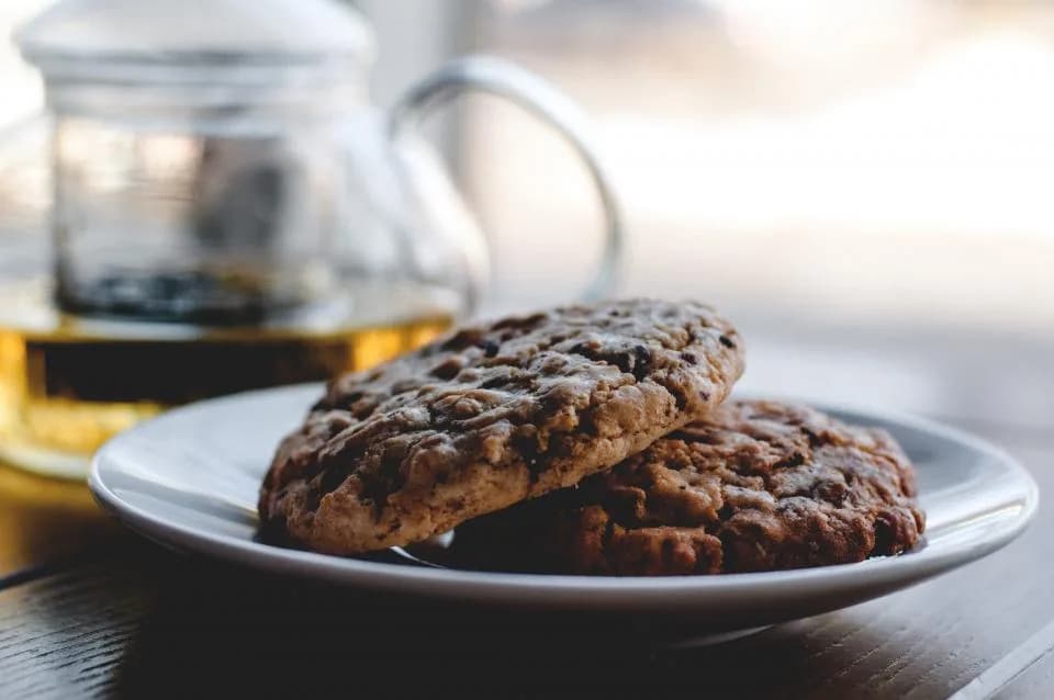 Dos galletas con trozos de chocolate en un plato, junto a una tetera